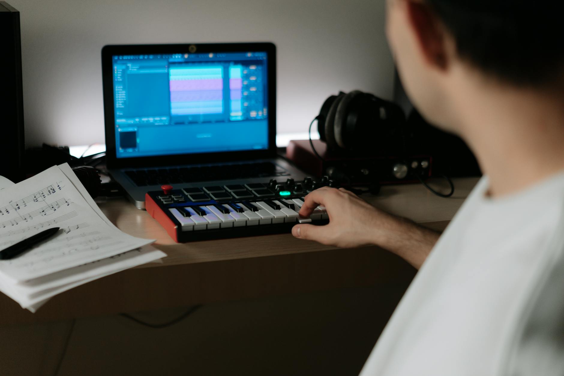 A music producer uses a synthesizer and laptop for composing in a home studio.